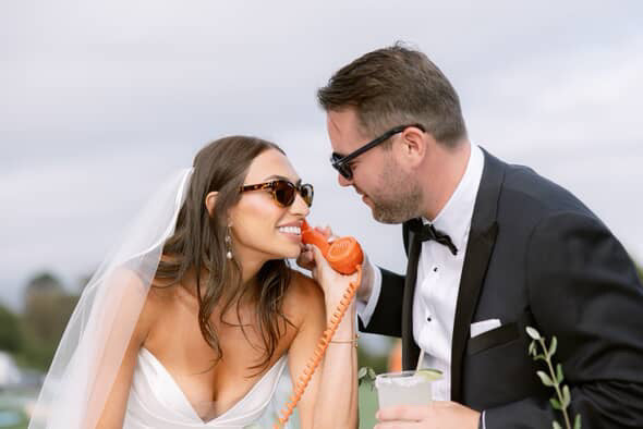 Bride and groom talk into rotary phone with sunglasses on