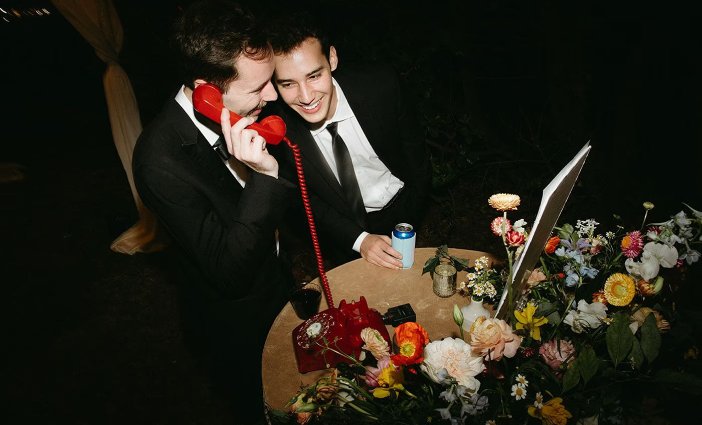 Two men in black tuxedos talking into red phone audio guestbook at wedding