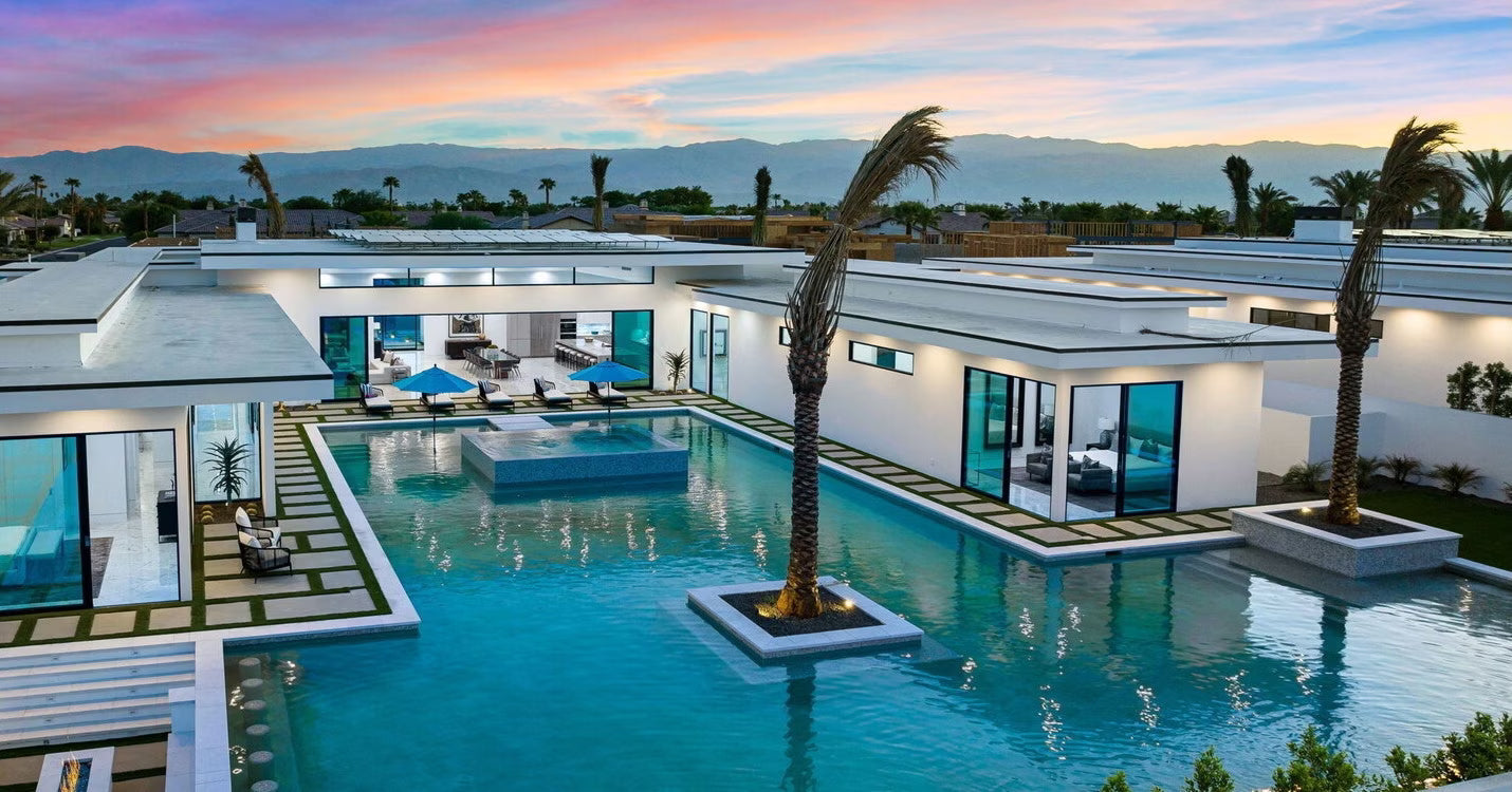 A modern desert estate with a stunning wraparound pool and mountain views at sunset.