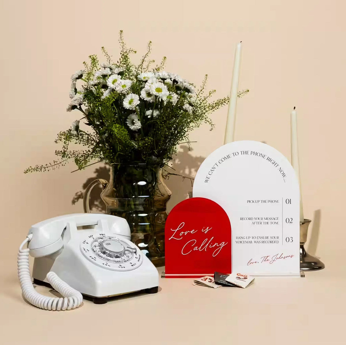 a white rotary phone next to a white and red sign and flowers with a beige backdrop