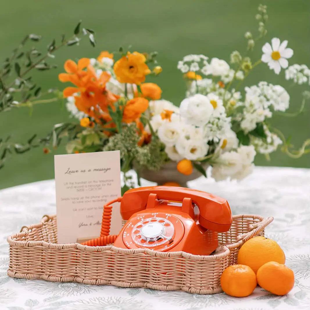 Vintage orange rotary phone in a woven basket with flowers and oranges on a light surface.