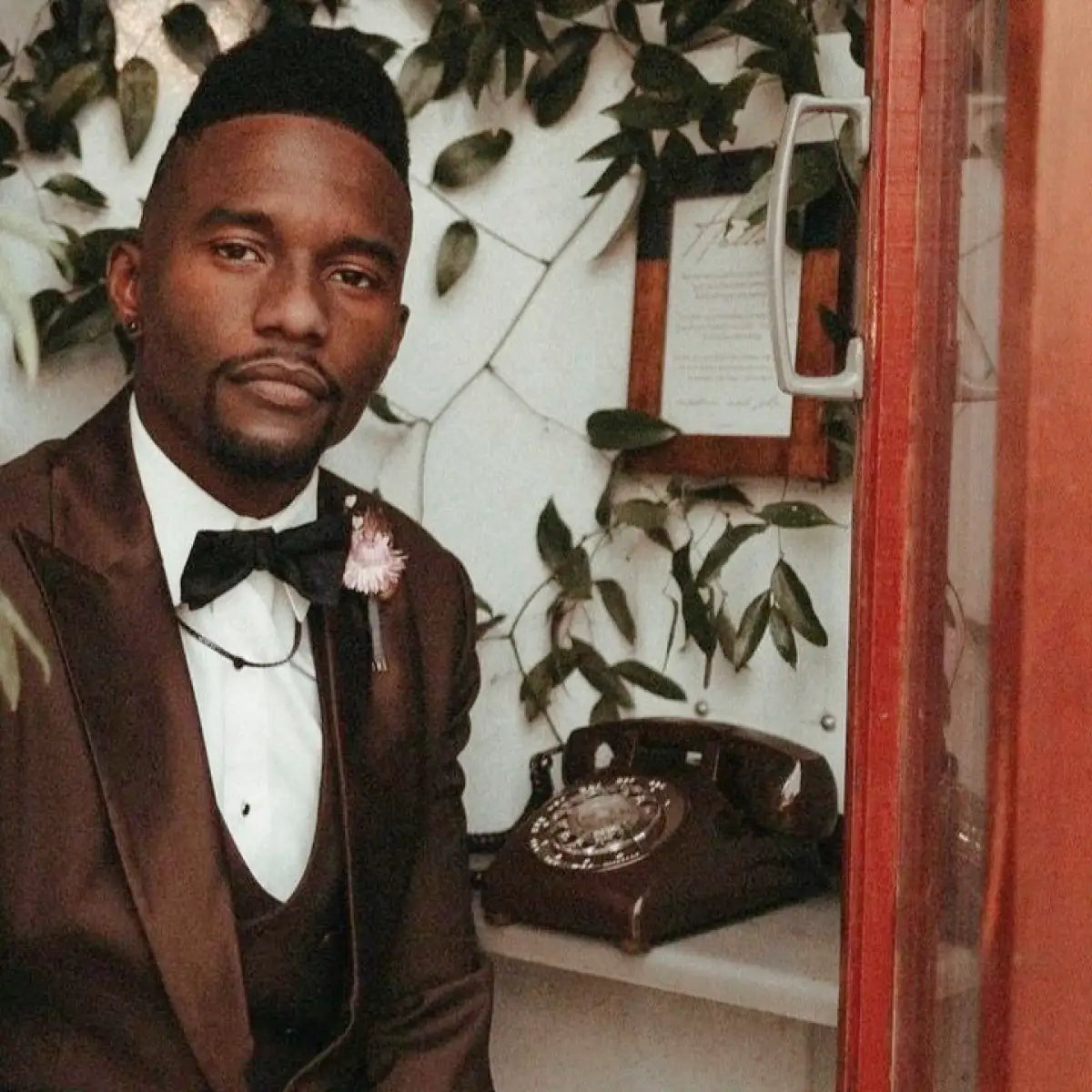 Man in a formal suit with a bow tie standing in a room with a brown vintage telephone and plants.