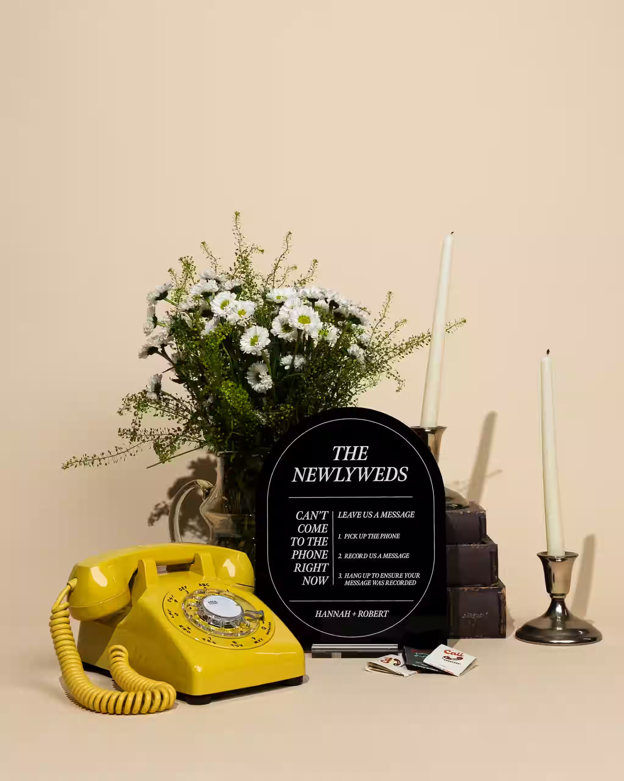 Yellow rotary phone, bouquet of flowers, and 'The Newlyweds' sign on a beige background