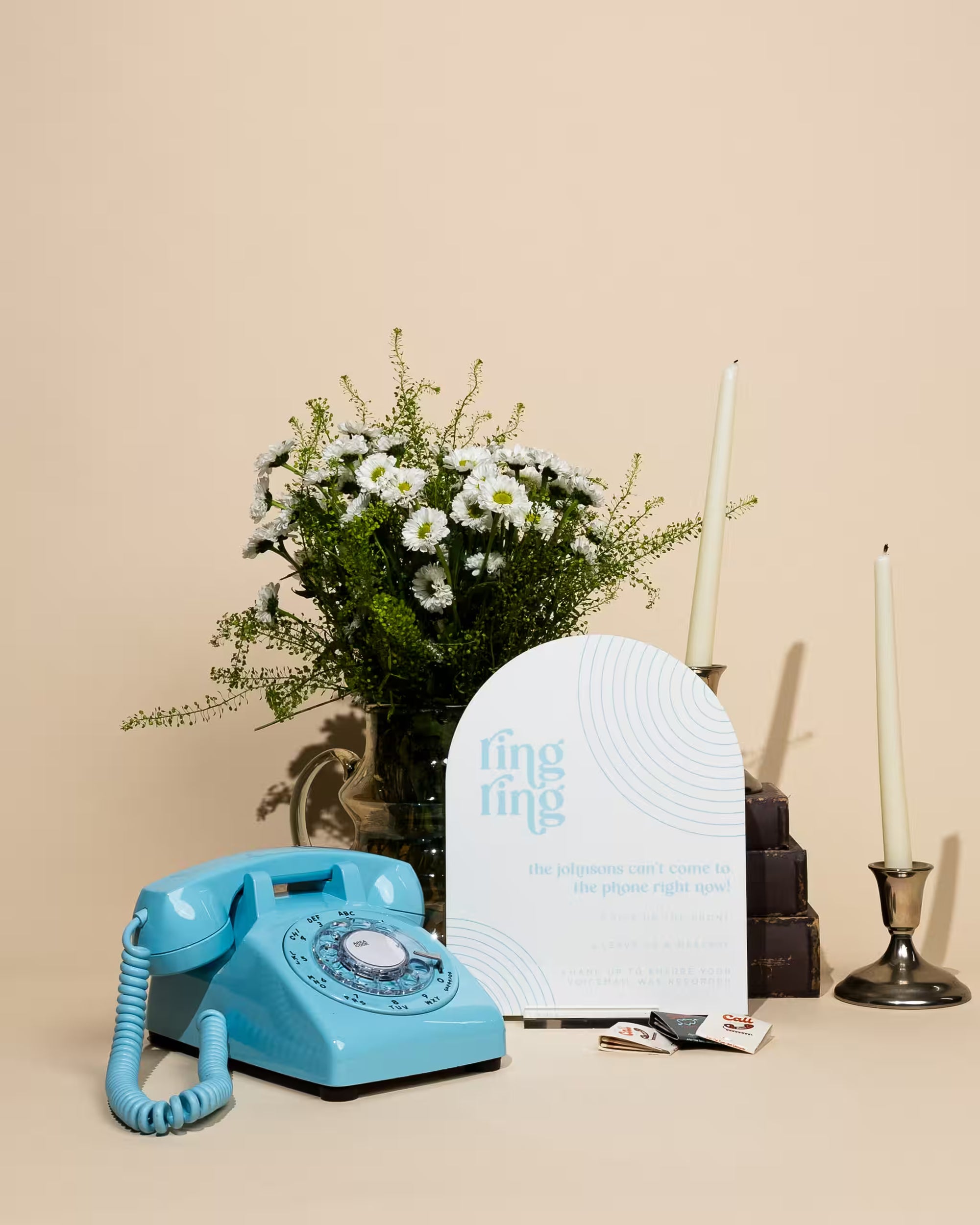 Vintage blue rotary phone next to a bouquet of flowers and a white arch-shaped object on a beige background
