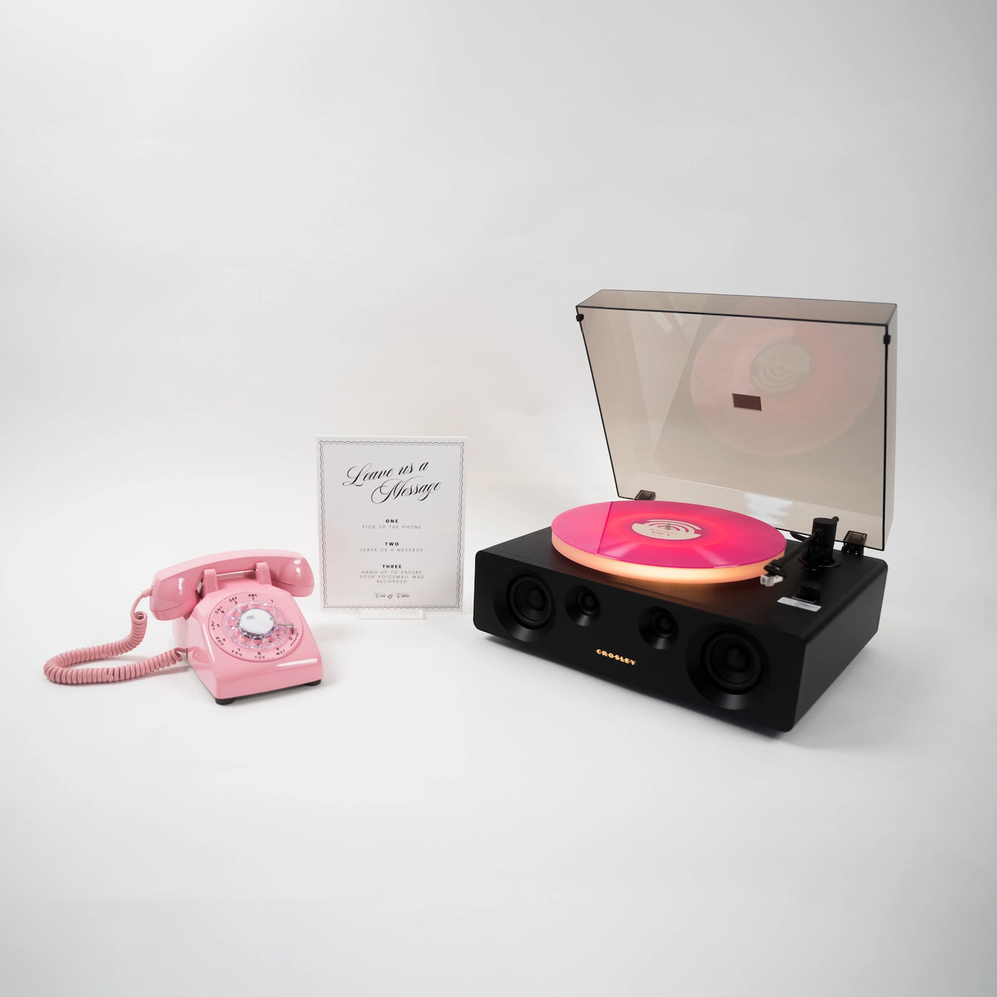 A retro-style black record player with a translucent dust cover open, showing a pink vinyl record on the platter, alongside a pink rotary telephone and a small framed card on a clean white background.