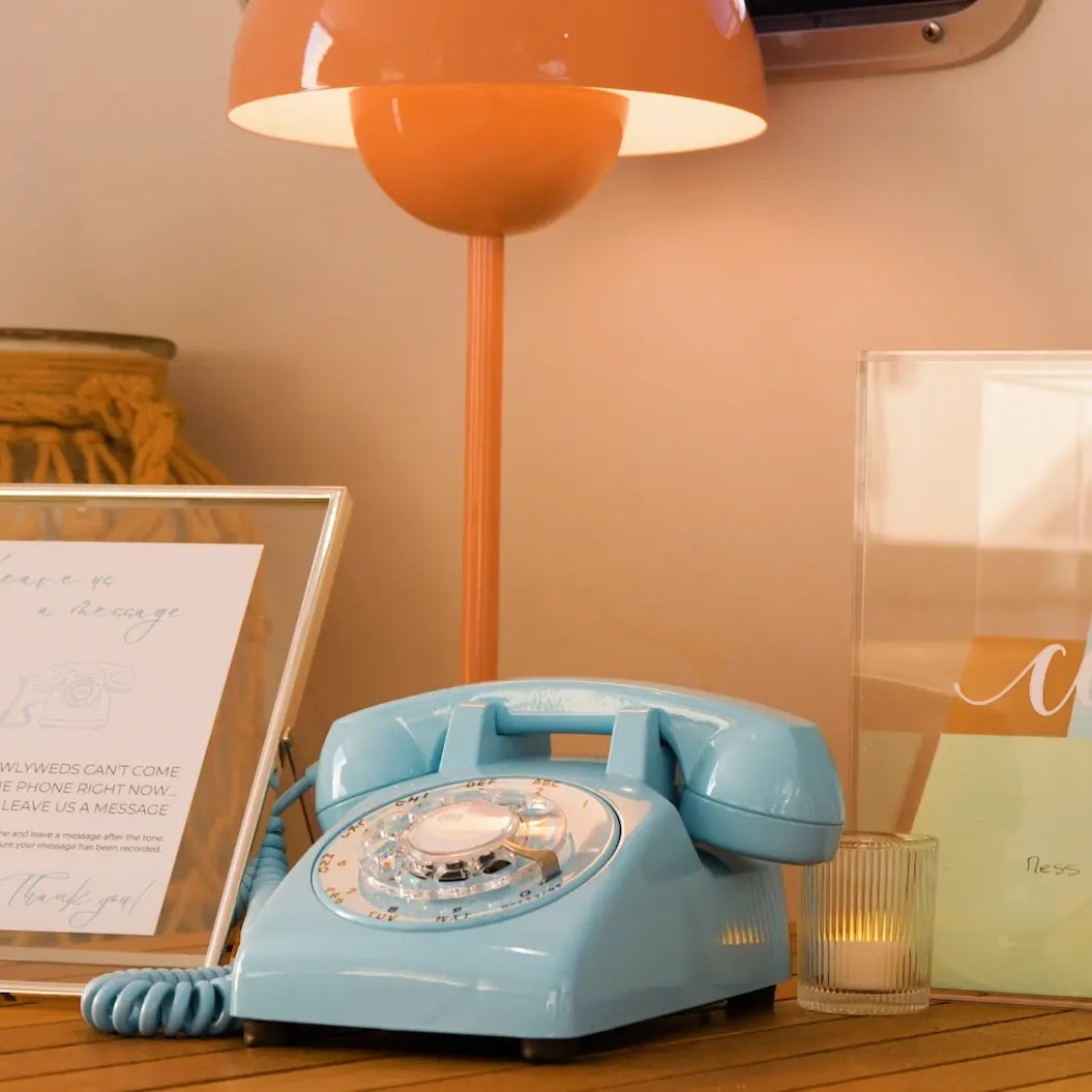 Vintage blue rotary phone on a wooden surface with an orange lamp and framed picture in the background.