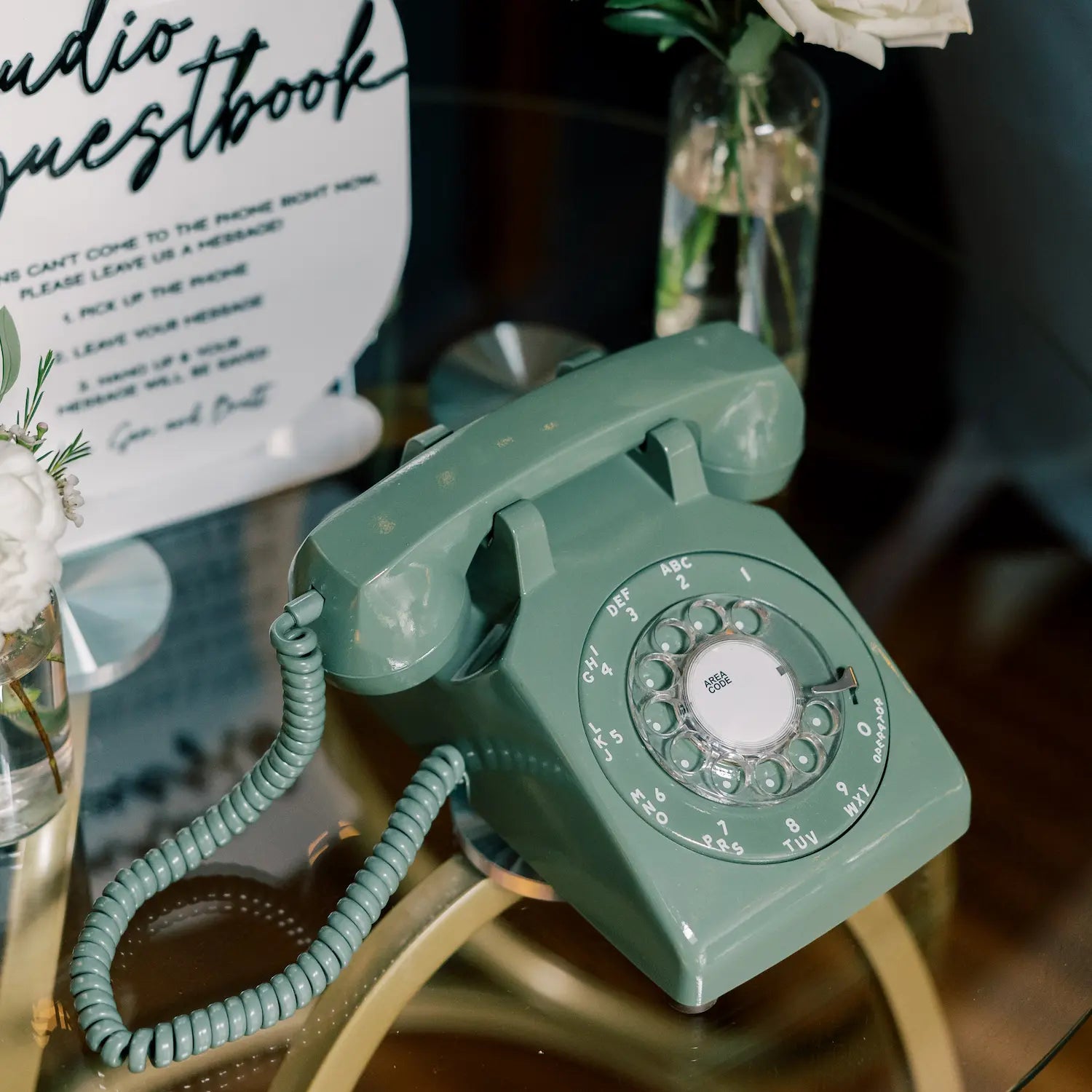 A green rotary phone on a glass table with a sign and flower behind it.