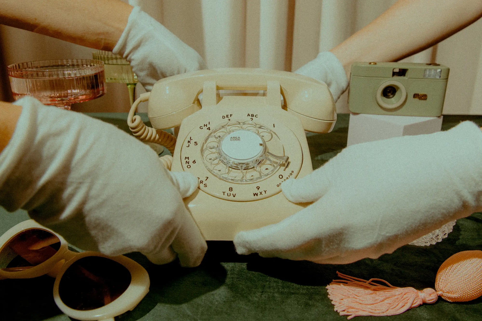 Vintage beige rotary phone held by gloved hands with a camera and other items in the background.