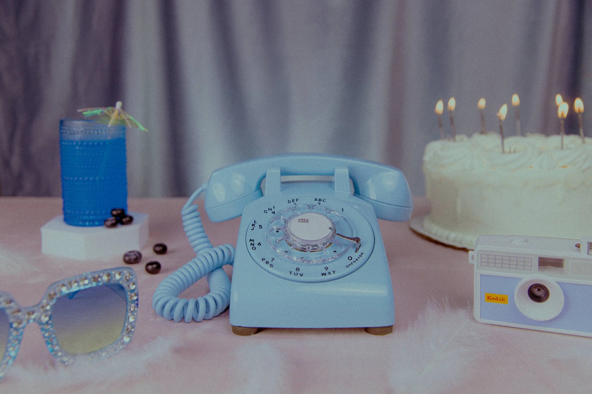 Vintage-style blue telephone on a table with a cake, sunglasses, and a drink.