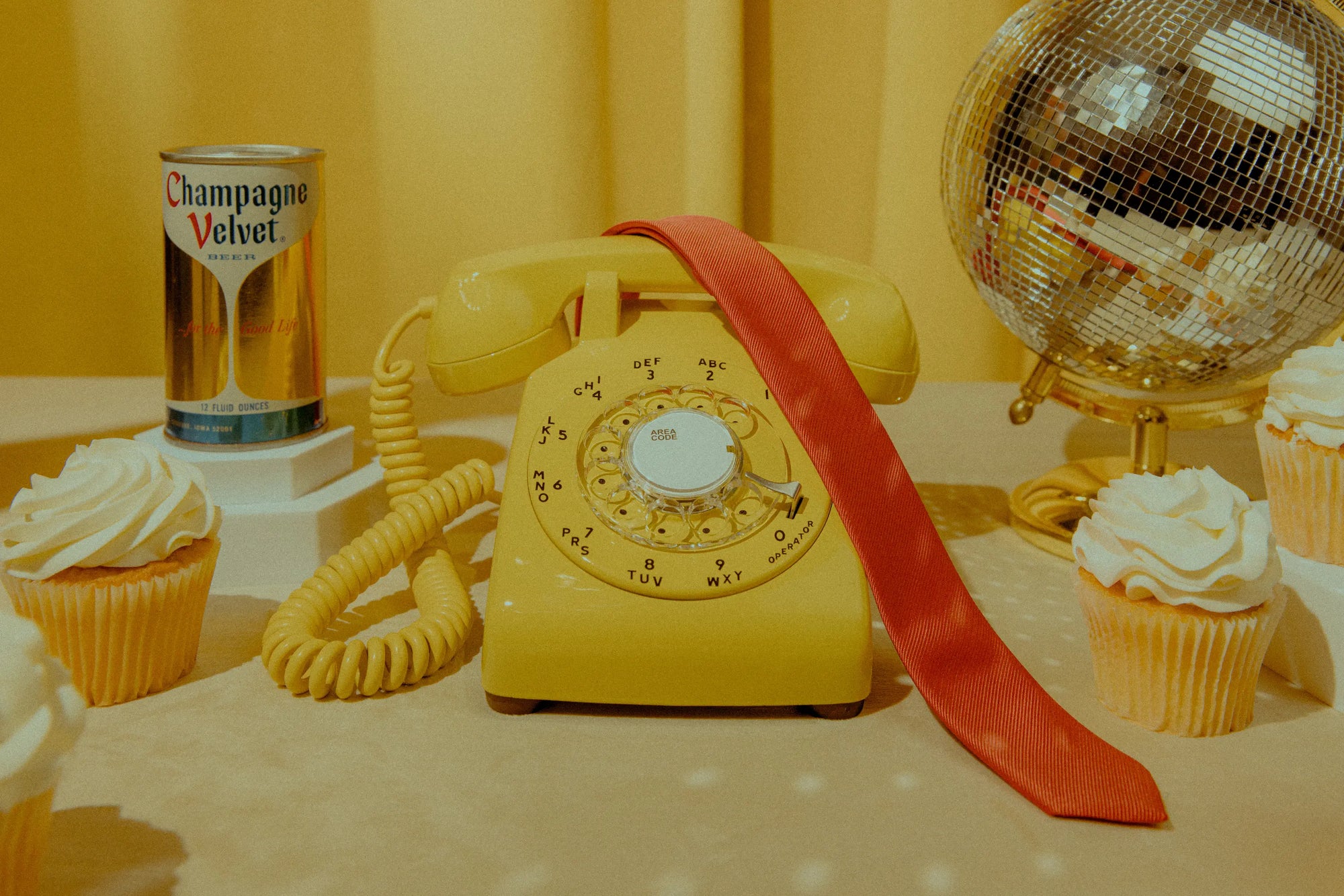 Yellow rotary phone with a red ribbon, beer can, and cupcakes on a yellow background
