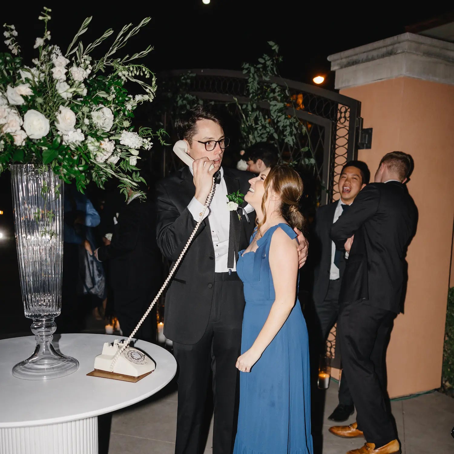 Man in formal attire with a toy phone, woman in blue dress, and other people in an outdoor evening setting.
