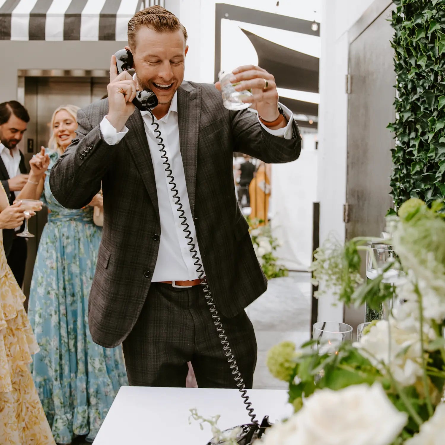 Man in a suit holding a phone and glass at an outdoor event with people and greenery.