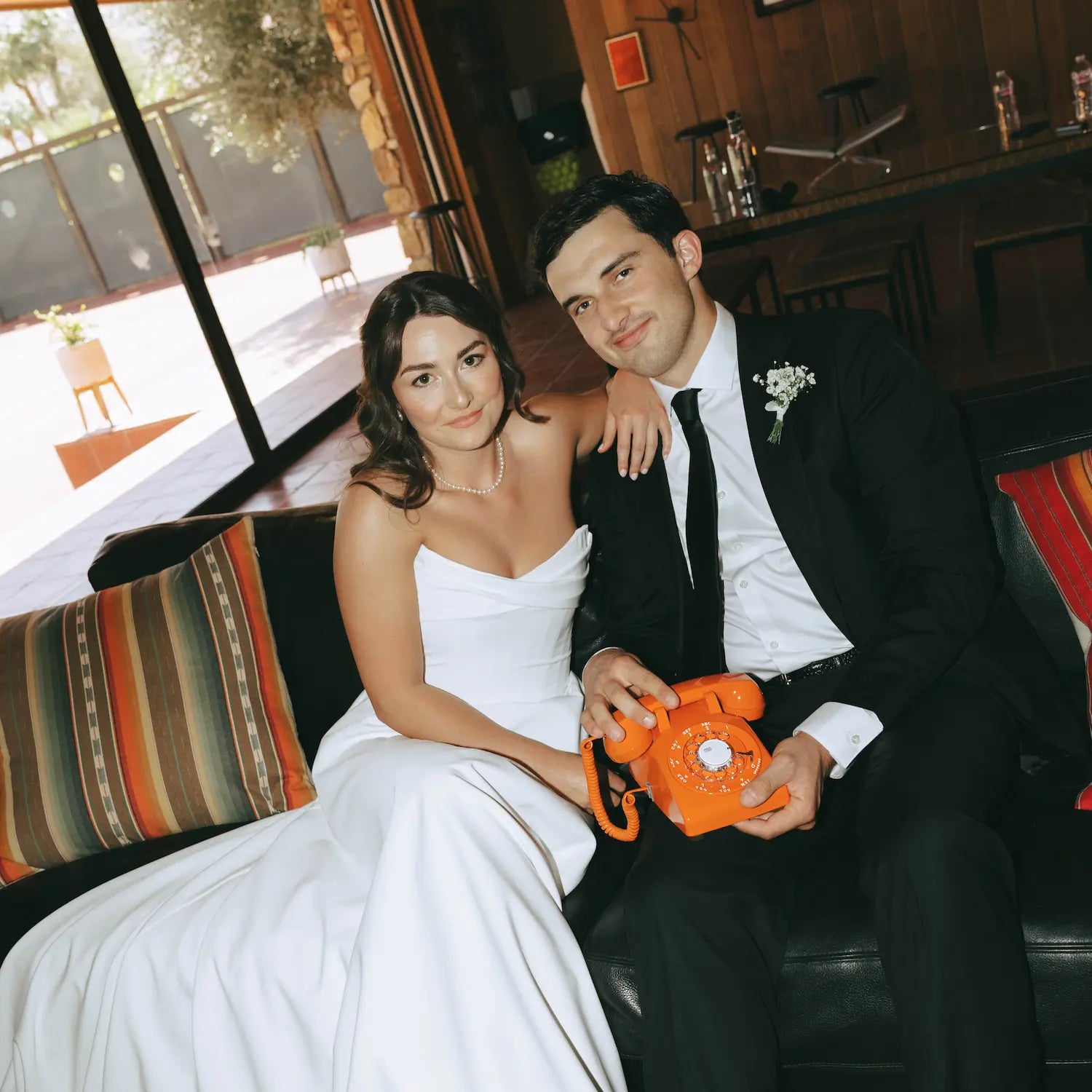 Man and woman in wedding attire holding an orange rotary phone in a casual setting.