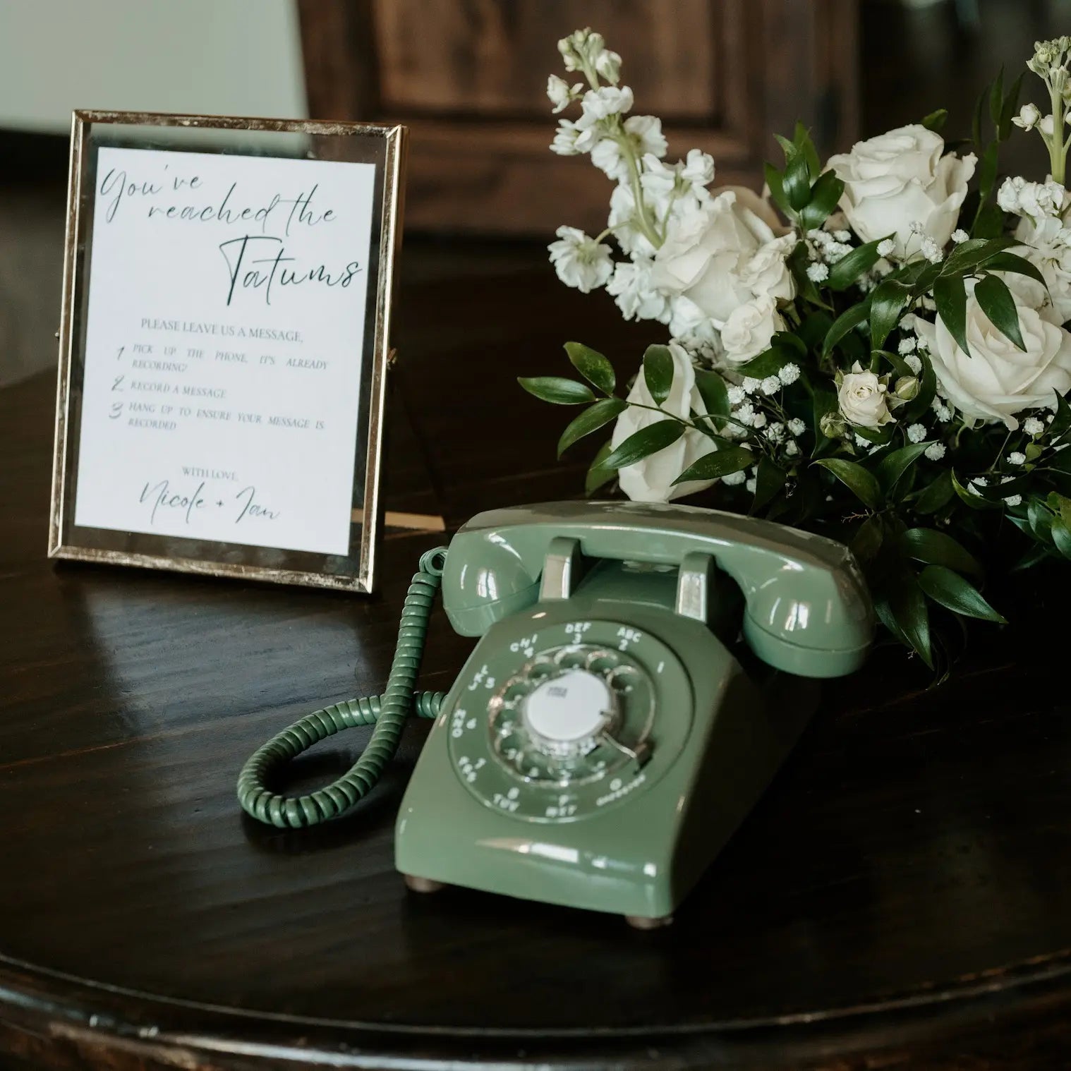 A green rotary phone with a sign and flowers behind it on a wood table.