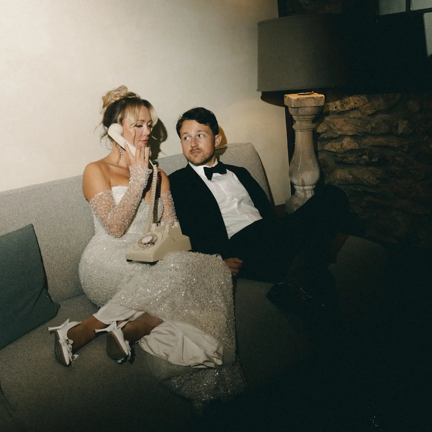 Man and woman in formal attire sitting on a couch in a dimly lit room using a cream rotary phone.