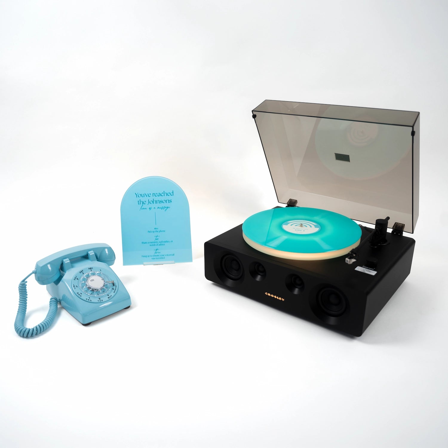 A novelty record player with a black body, a turquoise vinyl record, and an open transparent dust cover, shown beside a matching turquoise rotary telephone and a small blue card on a white studio background.