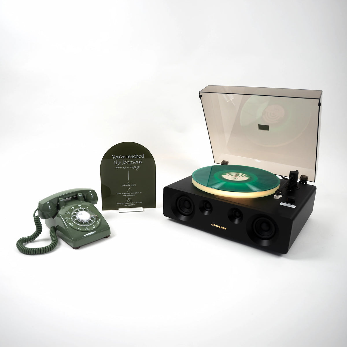 A black portable turntable with a translucent dust cover open, showing a green vinyl record, alongside a matching dark green rotary telephone and a small black plaque on a clean white background.