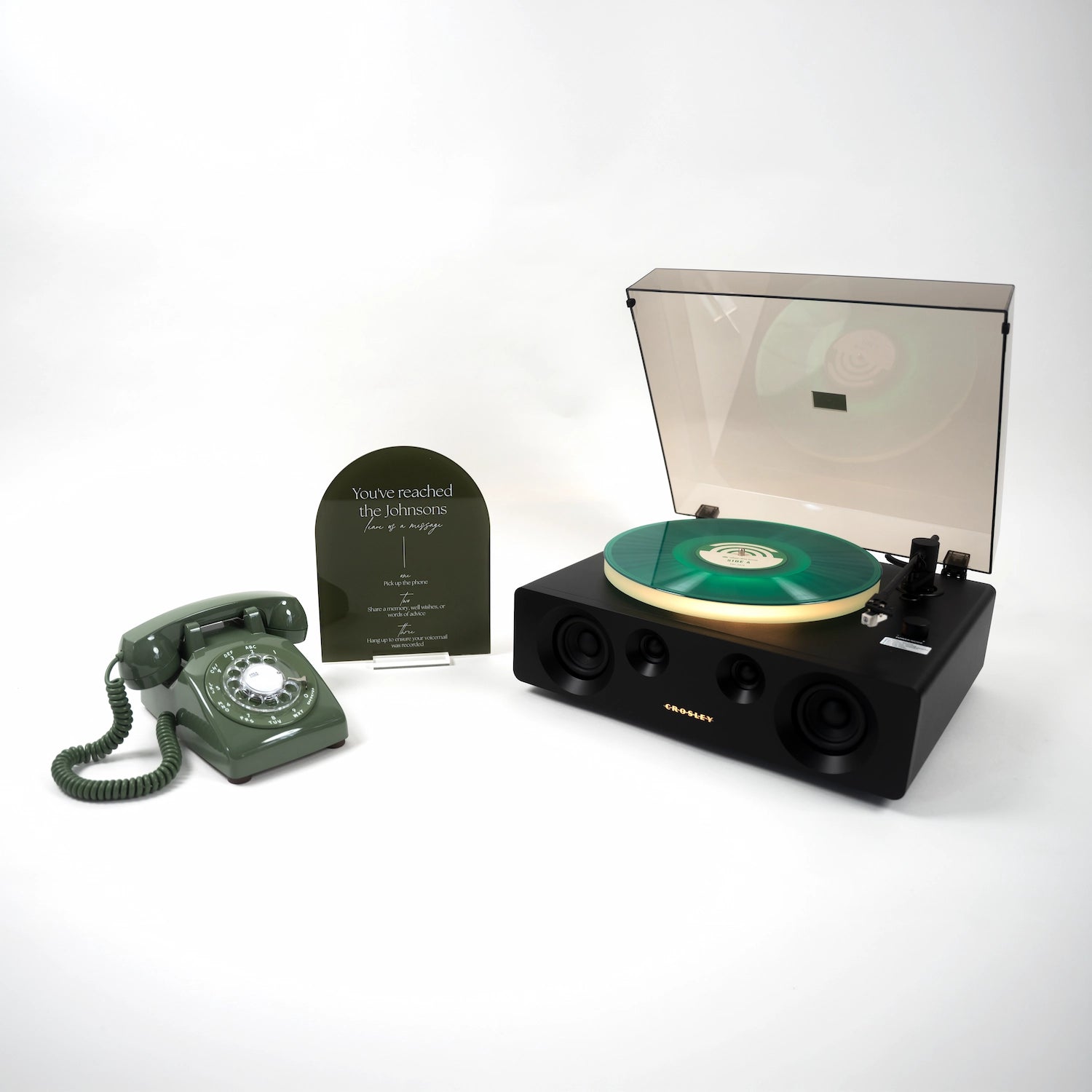 A black portable turntable with a translucent dust cover open, showing a green vinyl record, alongside a matching dark green rotary telephone and a small black plaque on a clean white background.