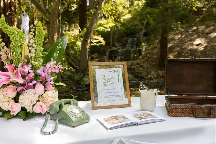 Decorative table setup with flowers, a vintage phone, and a sign in a forest setting