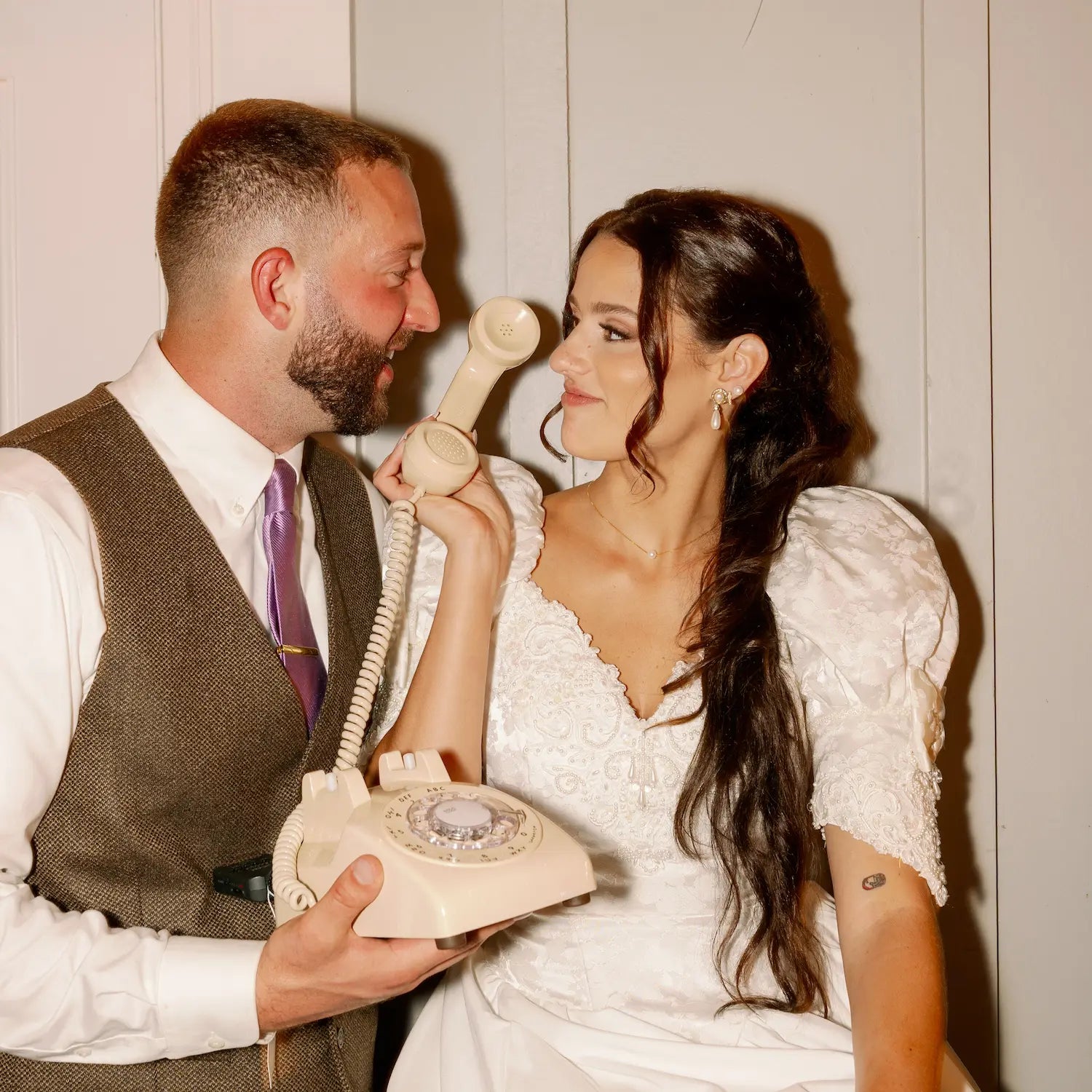 A man in a suit and a woman in a wedding dress using a cream audio guestbook rotary phone.