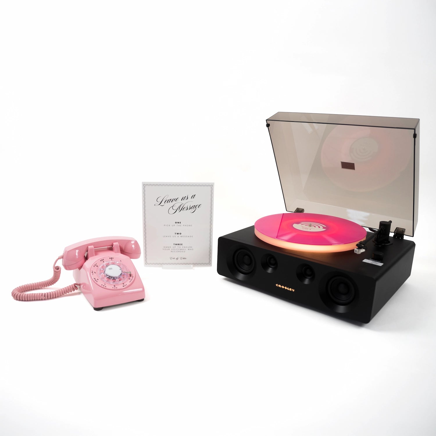 A novelty record player with a black body and a pink vinyl record, shown alongside a matching pink rotary telephone and a small card that reads 'Love is a Luxury'.