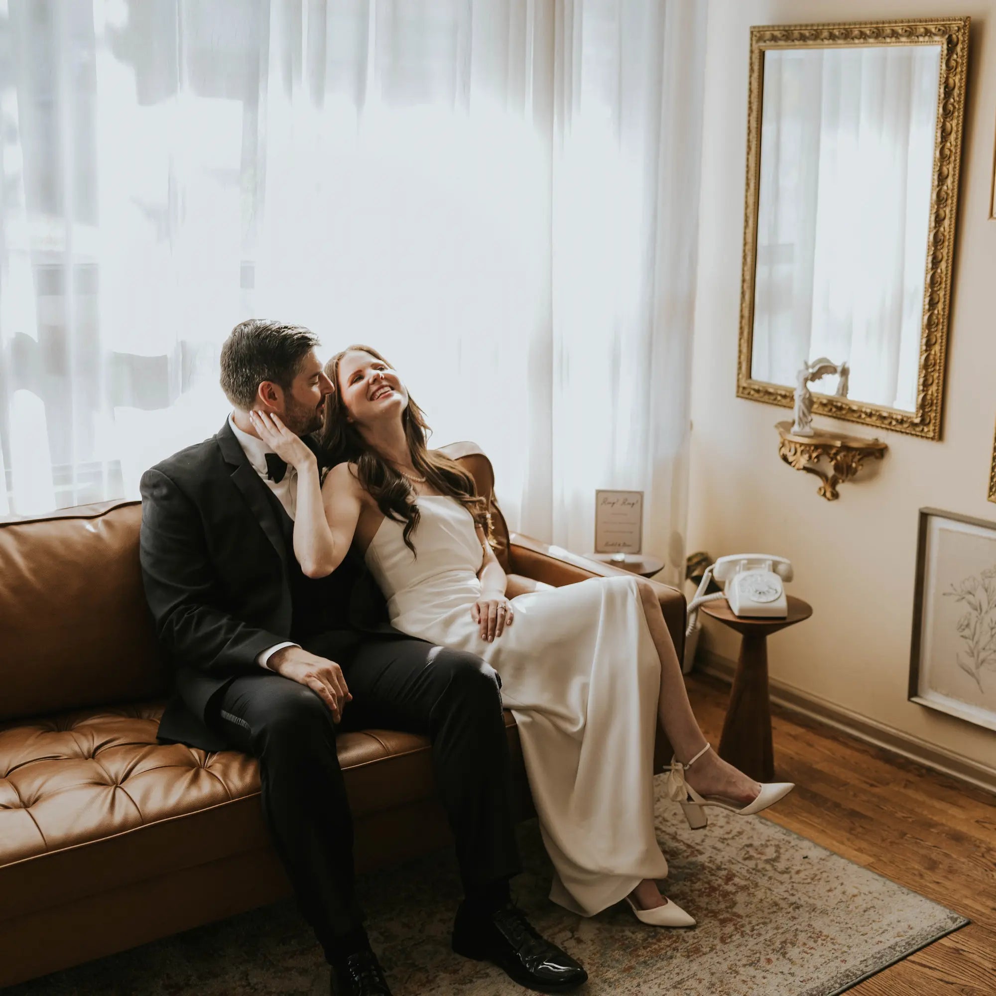 Couple sitting on a brown leather couch in a room with white curtains and a mirror.