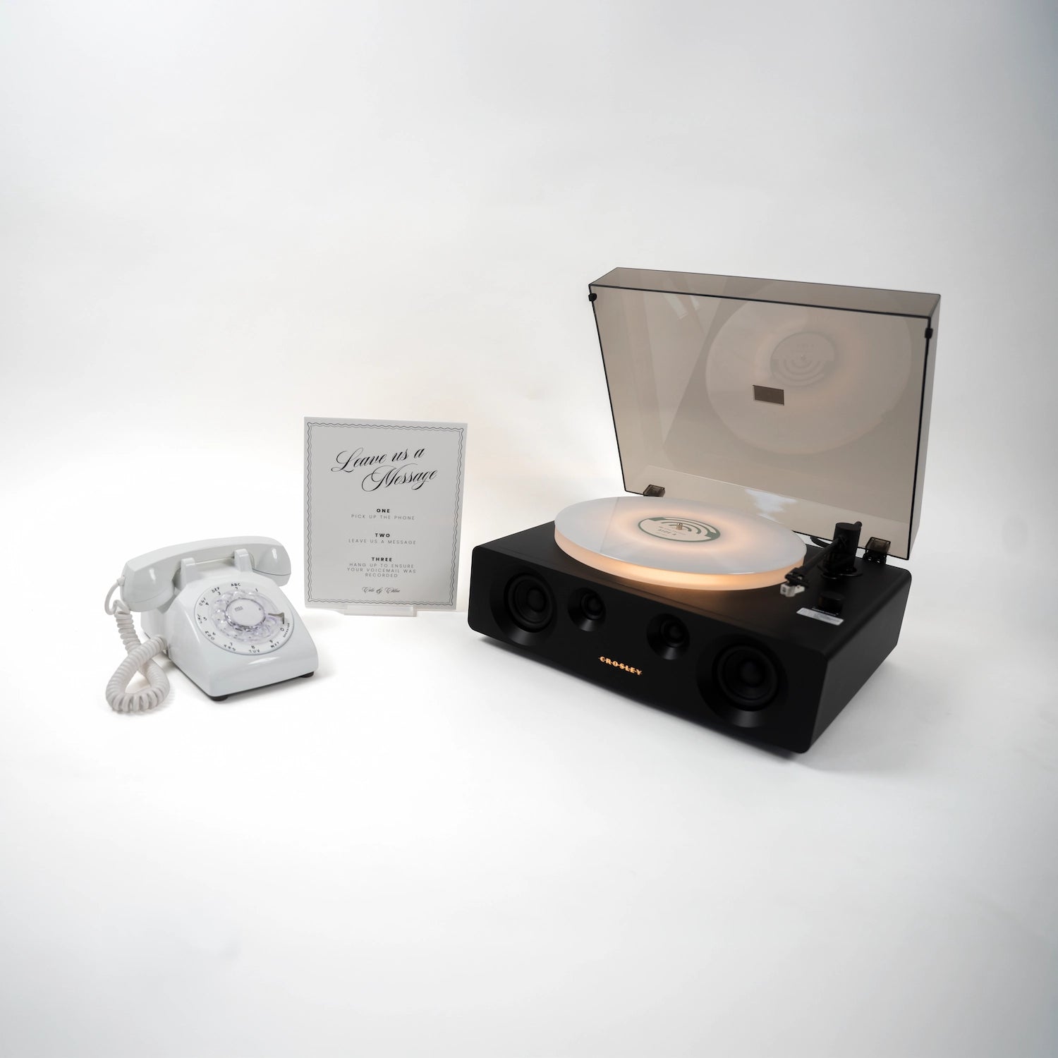 A retro-themed record player set featuring a black turntable with a translucent lid, a glowing vinyl record on the platter, a white rotary telephone, and a small framed sign on a clean white background.