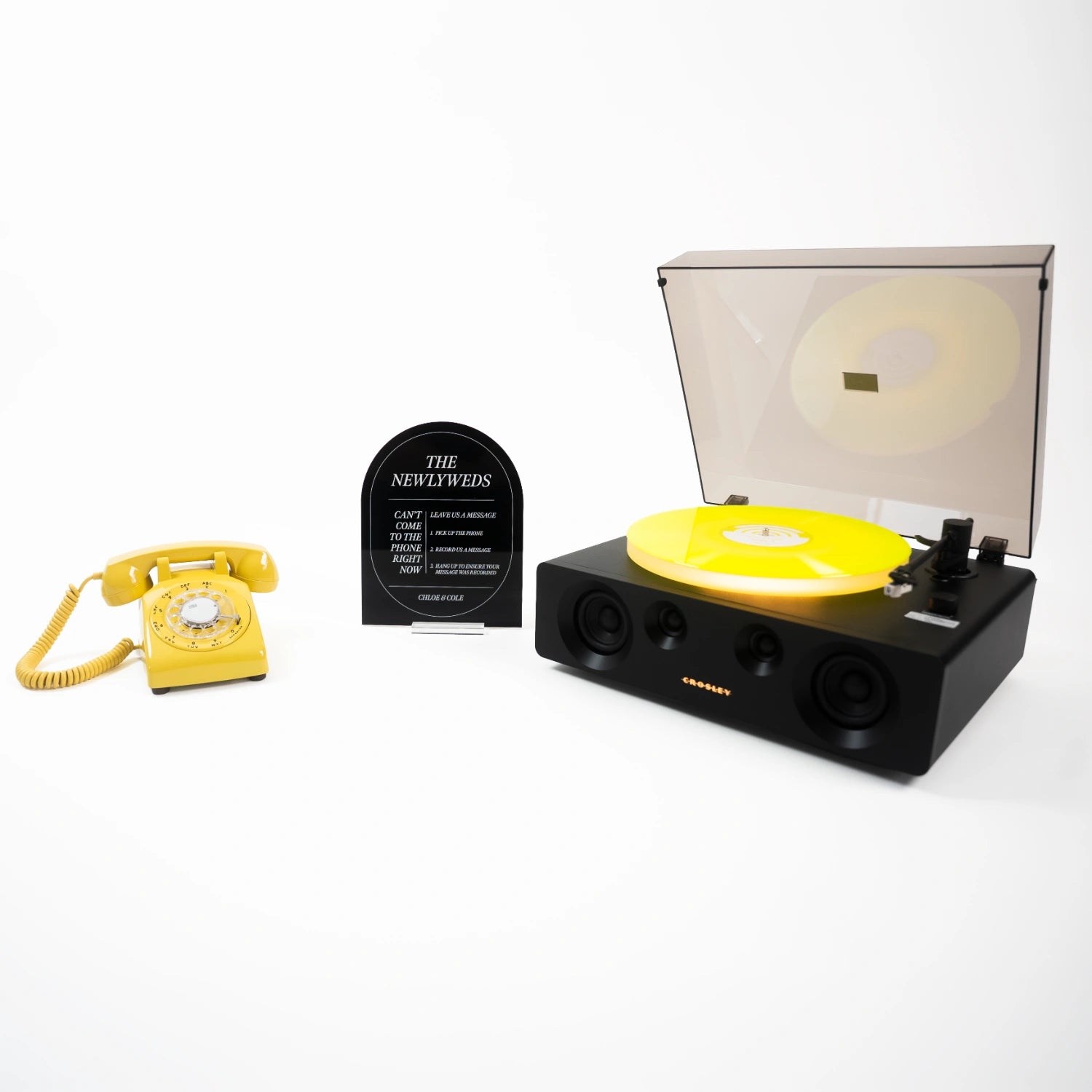 The Freddie bundle: a yellow rotary phone audio guestbook, a black Icon acrylic sign reading The Newlyweds, and a record player spinning a custom vinyl record.